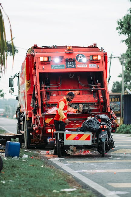 A street scene showing a red garbage truck with its rear compartment open, revealing mechanical components and a hydraulic system, parked on the side of a road. A worker wearing a high-visibility orange vest and dark trousers is standing in front of the truck, handling black plastic rubbish bags placed on a small metal platform attached to a scooter. The ground nearby is scattered with debris and litter, including a blue container on the grass verge. In the background, there are trees, utility poles, and distant commercial signage, with overcast sky and diffused natural light. The scene depicts a rubbish collection operation that may involve private waste disposal services, with the Rubbish Clearance Ruislip company possibly carrying out on-site clearance or waste removal from the local area.