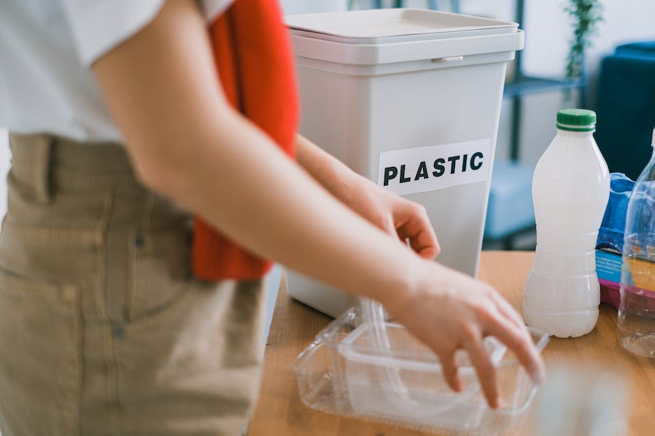 A person wearing a beige pair of trousers and a top with rolled-up sleeves is seen placing or retrieving a clear plastic food container from a designated recycling bin labeled 'PLASTIC' with bold black letters. The bin, made of light grey plastic with a flat hinged lid, is situated on a wooden surface or table. Around the bin, there are various plastic bottles, including a white one with a green cap, and other containers with colorful labels, indicating different types of recyclable plastic items. The setting appears to be an indoor environment, possibly a kitchen or recycling area, with soft lighting and a blurred background that suggests additional furniture or household objects. The scene emphasizes proper sorting of recyclable plastics, supporting the theme of independent rubbish disposal or waste separation, as handled by services like Rubbish Clearance Ruislip, and highlights the importance of accurate waste categorization for effective rubbish removal.