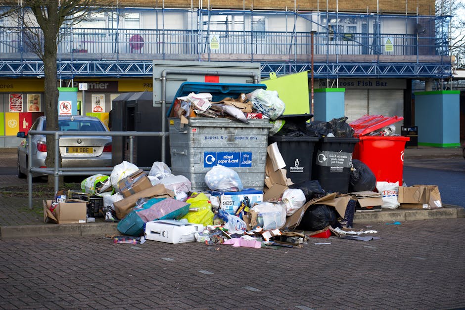 A street scene showing a red garbage truck with its rear compartment open, revealing mechanical components and a hydraulic system, parked on the side of a road. A worker wearing a high-visibility orange vest and dark trousers is standing in front of the truck, handling black plastic rubbish bags placed on a small metal platform attached to a scooter. The ground nearby is scattered with debris and litter, including a blue container on the grass verge. In the background, there are trees, utility poles, and distant commercial signage, with overcast sky and diffused natural light. The scene depicts a rubbish collection operation that may involve private waste disposal services, with the Rubbish Clearance Ruislip company possibly carrying out on-site clearance or waste removal from the local area.
