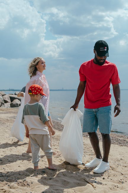 A group of three people, including a man and two children, are participating in a rubbish cleanup activity on a sandy beach near a body of water. The man, dressed in a red T-shirt, light blue denim shorts, and a black cap, is holding a large white garbage bag, appearing to collect litter. One child, wearing a beige and white outfit with a red polka-dotted cap, stands nearby holding another white bag, while a woman in a pink top observes from behind. The background features a partly cloudy sky with soft sunlight illuminating the scene, rocky shoreline on one side, and calm water extending into the distance. The setting emphasizes outdoor waste management and private disposal efforts consistent with alternative rubbish removal methods, with Rubbish Clearance Ruislip potentially facilitating such on-site cleanups away from local authority collection.