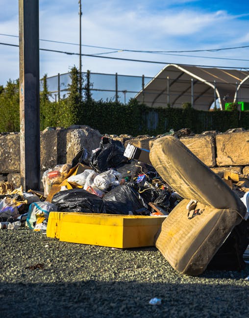 A large collection of rubbish and waste materials piled on a paved urban street corner, including black and red plastic trash bags, flattened cardboard boxes, paper and plastic packaging, and discarded miscellaneous items. The waste is situated near a metal railing separating a parking area, with a silver car parked on the left side. Behind the waste, there are commercial storefronts with yellow signage displaying emergency and customer information, and a building under construction or renovation, covered with blue scaffolding and safety netting. The scene is lit with natural daylight, providing clear visibility of the various textures and colors of the waste—ranging from shiny plastic bags to rough cardboard surfaces—and the surrounding urban environment. This depiction exemplifies a typical scenario where private waste collection or rubbish clearance services, such as those by Rubbish Clearance Ruislip, are required to manage and thoroughly clear accumulated rubbish in local streets and public spaces.